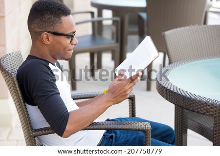 Closeup portrait, smart young nerdy man with big black glasses, sitting down and reading, isolated outdoors background.  knowledge is power concept