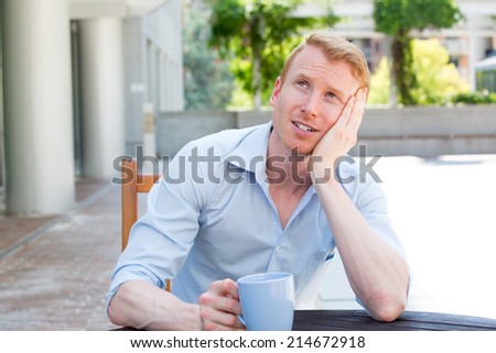 Closeup portrait, young business man drinking mug outside, sitting on wooden chair, worrying, daydreaming, isolated outdoors, outside background