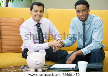 Closeup portrait, two young men in ties shaking hands, isolated yellow couch background with piggy bank and coffee on table foreground