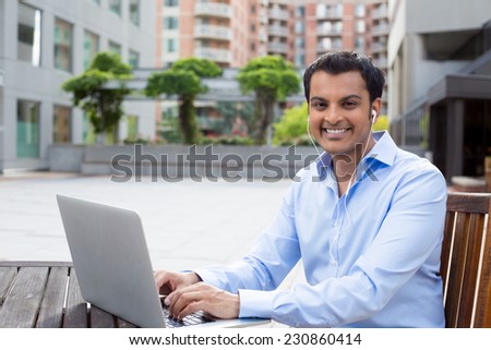 Closeup portrait, happy young handsome man in blue shirt typing away, browsing digital computer laptop, isolated background of sunny outdoor, green trees, office background