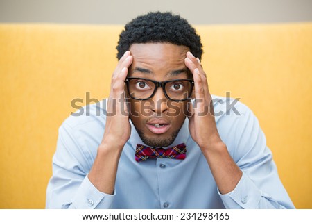 Closeup portrait, stunned nerd young man, hands on head, open mouth jaw drop with bow tie and big glasses, isolated yellow background. Negative human emotion facial expression feelings