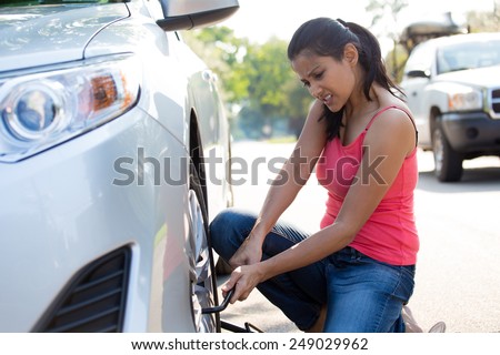 Closeup portrait, young woman in pink tanktop and blue jeans fixing flat tire with jack and tire iron, isolated green trees and road outside background. Roadside assistance concept