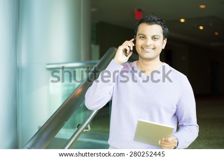 Closeup portrait, handsome young business man, happy guy, in purple sweater, using cell phone and tablet, smiling, having pleasant conversation, isolated indoors office. Human emotions, expression