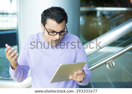 Closeup portrait, young angry man in black eyeglasses and purple sweater, astonished, wide open mouth, by what he sees on tablet, isolated indoors office background
