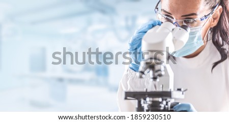 Female lab technician in protective glasses, gloves and face mask sits next to a microscope in laboratory.
