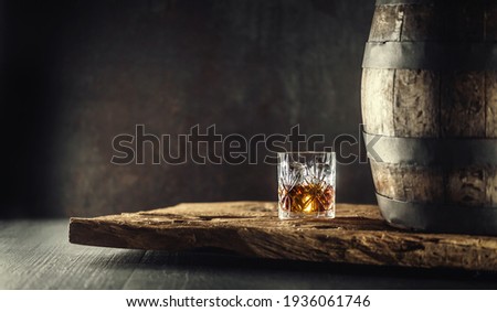 Glass of whisky cognac or bourbon in ornamental glass next to a vinatge wooden barrel on a rustic wood and dark background.