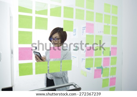 Young Vietnamese manager with stack of document using smart phone on the background of sticky-notes wall