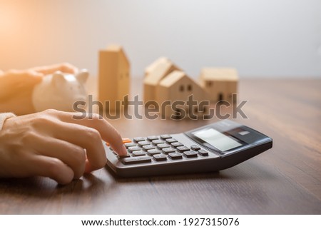 The hand is pressing calculators, piggy bank with wooden house. buy or rent question on note with calculators on desk. Save money and buy house concept.
