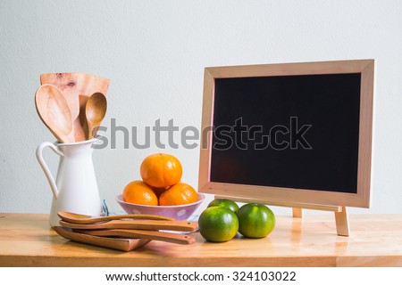 Kitchenware with wooden ladle, wooden spoon in white vase, blackboard and orange on wooden table over white wall background