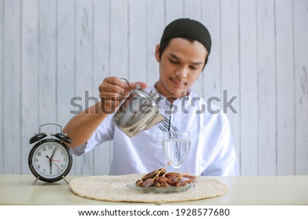 Muslim man wearing koko pouring water into a glass on the table, preparation for iftar. Traditional Ramadan, iftar meal. Ramadan kareem fasting month concept. Selective focus