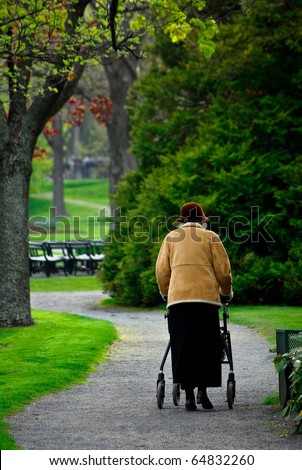 An elderly lady takes a walk alon a park path. she is wearing a hat brown coat and skirt and is using a walker