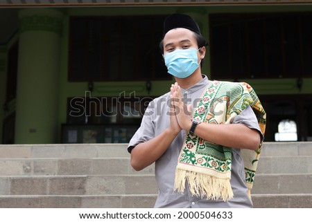 Portrait of religious Asian man in koko shirt with prayer mat slung over the shoulder, wearing medical mask, showing apologize and welcome hand gesture. Outdoor shoot in front of mosque Imagine de stoc © 