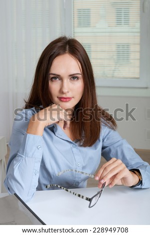 Girl sits at a table in office holding glasses and propped her head with her hand