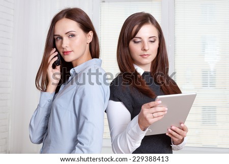 Two women in formal clothes standing back to back. One calls on the phone and the other reads the information in her tablet