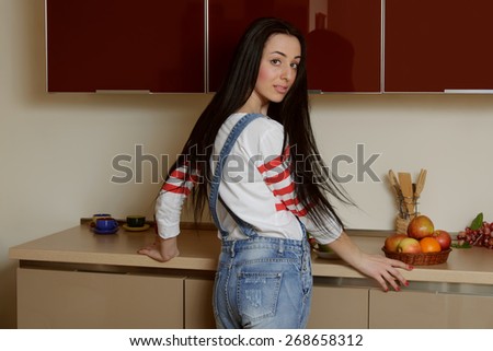 Brunette girl in home clothes standing near a kitchen cupboard in half a turn. She is dressed in blue overalls and a white shirt. Behind her is a kitchen furniture painted in beige and maroon tones.