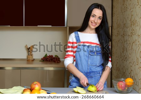 Brunette housewife in blue denim overalls cut green apple with a knife. The girl looks at the camera and smiling. She is standing in the kitchen painted in beige tones. On the table are fruits.