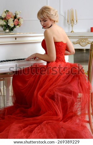 Woman dressed in a red evening gown with a long train sits and playing on a white piano looking slightly to the side against a light background