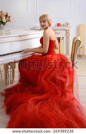 Female dressed in a red evening gown with a long train sits and plays on a white grand piano looking slightly to the side against a light background