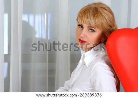 Portrait of business woman in formal attire who sits in a red chair in a half-turn against the light background