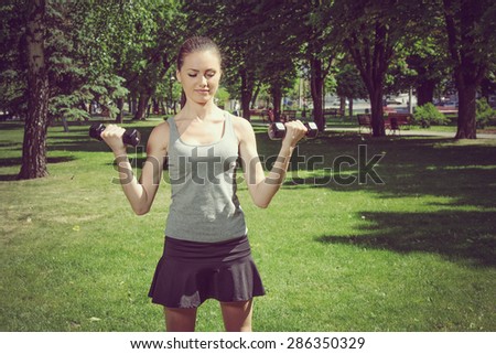 Activities girl doing exercise with light dumbbells standing on the green grass in the street. Healthy lifestyle concept.