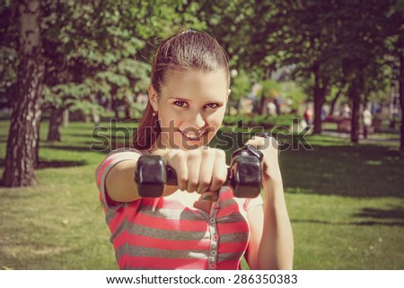 Fitness woman simulates a hit with light dumbbells in the park outdoors