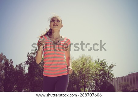 Sport woman in red t-shirt running and listening to music with headphones in the phone outdoors against the sky