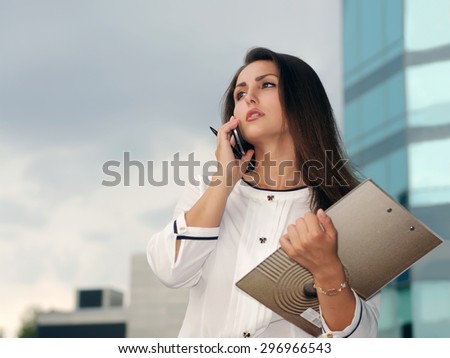 Business woman in formal clothes is on the city\'s business district with a folder in her hand and talking on the phone