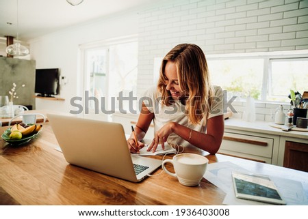Caucasian female student working from home writing in note pad with laptop sitting next to hot coffee