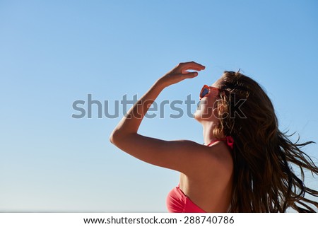 Attractive young woman with fit healthy body wearing bikini tying up hair low angle shot with blue sky background showing tropical vacation concept