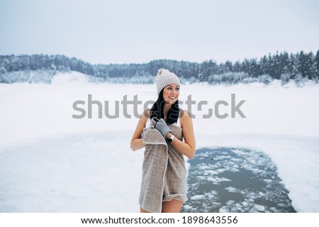 Winter swimming. Woman ready to swim in ice water. How to swim in cold water. Beautiful young woman wrapped in a towel and swimming clothes. Gray hat and gloves. People and nature lake in the forest.