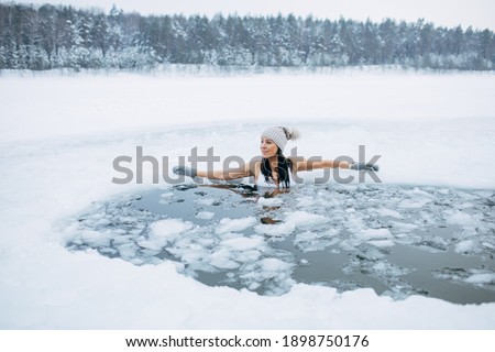Winter swimming. Woman in frozen lake ice hole. Swimmers wellness in icy water. How to swim in cold water. Beautiful young female smiling. Gray hat and gloves swimming clothes. Nature lake in forest