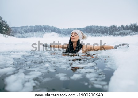 Winter swimming. Woman in frozen lake ice hole. Swimmers wellness in icy water. How to swim in cold water. Beautiful young female in zen meditation. Gray hat and gloves swimming clothes. Nature lake