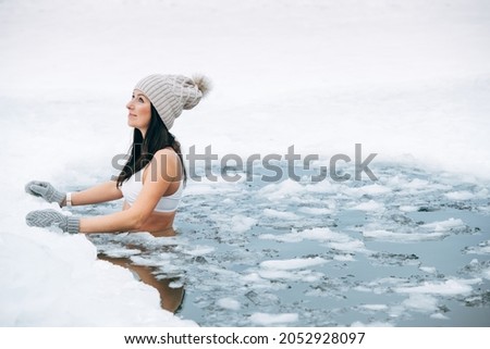 Woman in frozen lake ice hole. Winter cold swimming. Swimmers wellness in icy water. How to swim in cold water. Beautiful young female in zen meditation. Hat and gloves swimming clothes. Nature lake