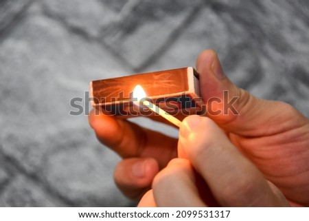 Detail of ignition of matches for a matchbox. A burning match in one hand and a matchbox in the other. The process of lighting a match. Hand with burning match on gray background