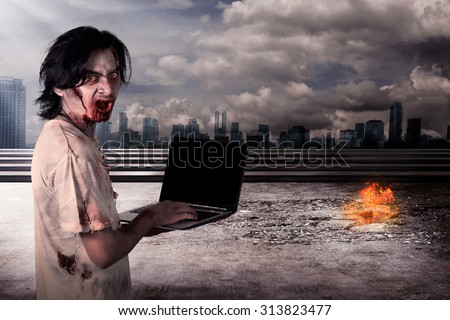 Creepy male zombie typing with laptop with city on fire background