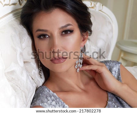 Sersualnaya beautiful young brunette woman with evening make-up chic groomed wearing a short evening dress embroidered with silver sequins and earrings sitting on the couch in background with flower