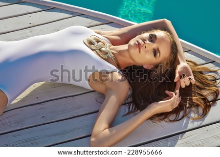Beautiful blonde girl in good shape with long light hair and tan perfect skin in white swimming suite with golden jewelry with lie near the pool with green water keeping open her eyes with soft smile