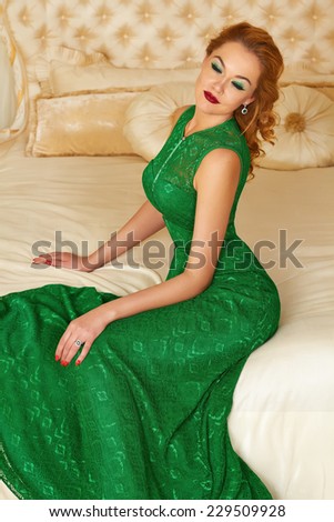 Beautiful young red-haired curly woman with bright makeup crimson lipstick, wearing a long bright designer evening dress, green earrings with emerald and  diamonds, sitting on bed in  bedroom interior