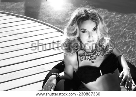 Black and white photo of beautiful sexy blonde woman laying near the swimming pool in black suite and bracelet and colorful beads