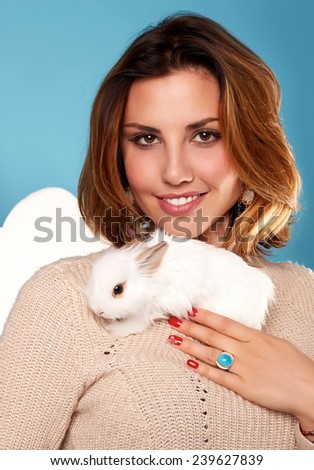 Beautiful young girl with blond hair bright with natural make-up smiling red manicure wearing beige warm sweater is holding the white fluffy little rabbits on a background of blue sky and white clouds