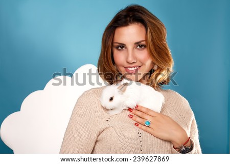Beautiful young girl with blond hair bright with natural make-up smiling red manicure wearing beige warm sweater is holding the white fluffy little rabbits on a background of blue sky and white clouds