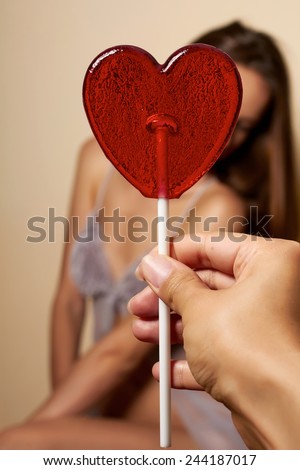 Beautiful young girl in a dressing gown of silk and lace with thin spaghetti straps with a cut and bow in front of hands holding lollipop in the shape of a red heart looking into camera valentines day