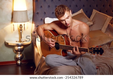 Handsome young sexy man with a naked torso muscular tanned body athletic figure sitting in the bedroom lamps veserom at playing a musical instrument guitar romance