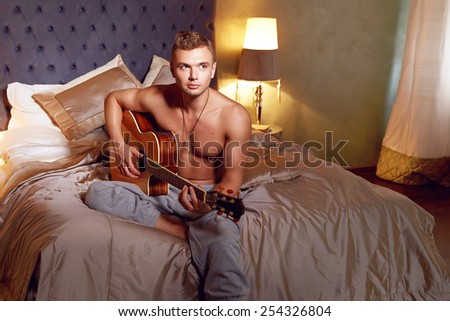 Handsome young sexy man with a naked torso muscular tanned body athletic figure sitting in the bedroom lamps veserom at playing a musical instrument guitar romance
