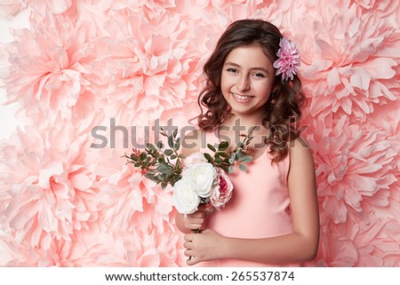 Beautiful young little girl with long wavy brunette hair with a bright evening make-up perfect summer tan thin figure dressed in colored short dress holding flower pink