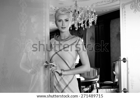 Beautiful young sexy blonde with short hair exquisite evening make-up standing at the door an interior in fashionable stylish dress bare shoulders lamp sofa Baroque
