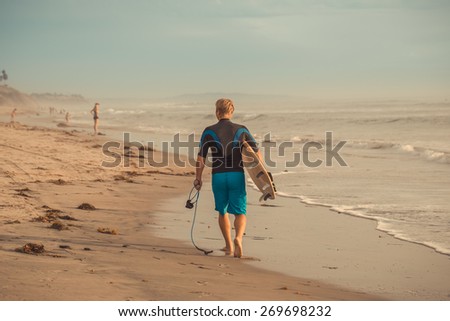 Man surfer walking along the beach with surfboard at sunset.