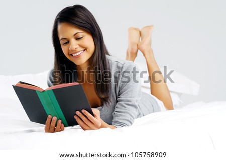 Woman reading a book in bed, lying on her stomach smiling happy and relaxed on a leisure day at home.