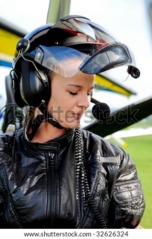 A girl poses with flight gear. she wears a helmet, radio equipment including a microphone, headset and helmet. she wears a black flight suit.