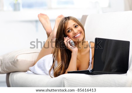 Woman lying on the couch using her laptop and having a happy conversation on the telephone